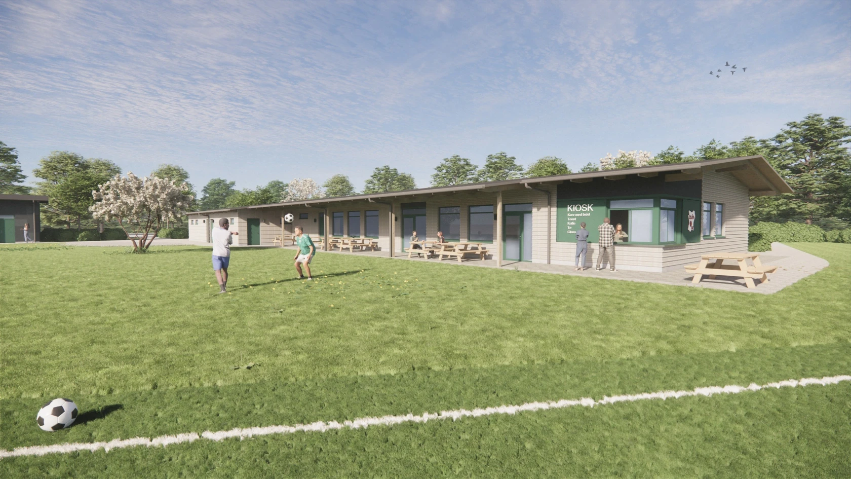 Fotbollsplan med barn som leker med boll, i bakgrunden syns en enplans byggnad med bänkar utanför och en litet kiosk