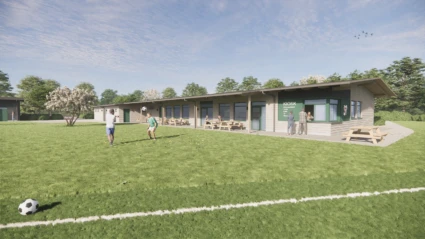Fotbollsplan med barn som leker med boll, i bakgrunden syns en enplans byggnad med bänkar utanför och en litet kiosk