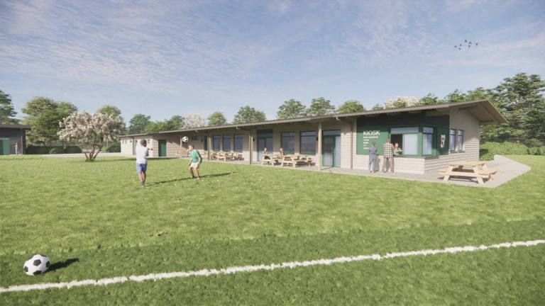 Fotbollsplan med barn som leker med boll, i bakgrunden syns en enplans byggnad med bänkar utanför och en litet kiosk